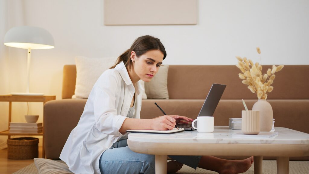 Mulher jovem aplica aprendizagens sobre como ser mais disciplinada enquanto tira apontamentos em frente a um laptop para estudar para um exame.