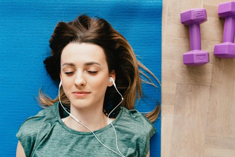 Jovem mulher deitada num tapete de yoga, com headphones nos ouvidos põe em prática técnicas de relaxamento como a meditação e o mindfulness.