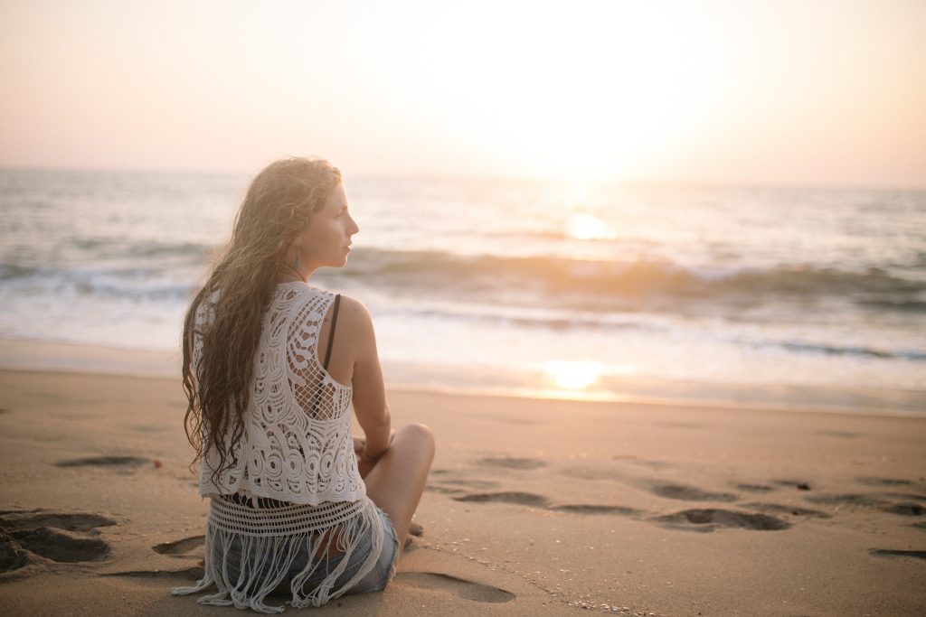 Mulher, sentada à beira-mar ao pôr do sol, observa o horizonte numa prática ativa de meditação e mindfulness.
