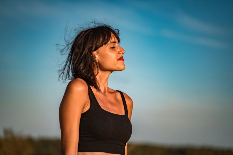 Jovem mulher ao ar livre usufrui dos benefícios da meditação numa prática de mindfulness, sentindo o vento e o sol na pele.