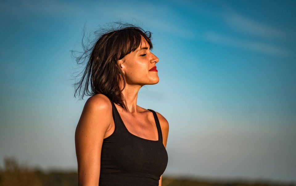 Jovem mulher ao ar livre usufrui dos benefícios da meditação numa prática de mindfulness, sentindo o vento e o sol na pele.